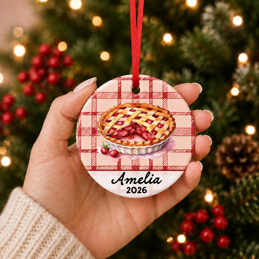 Hand holding personalized cherry pie ceramic ornament with name and year beside a Christmas tree
