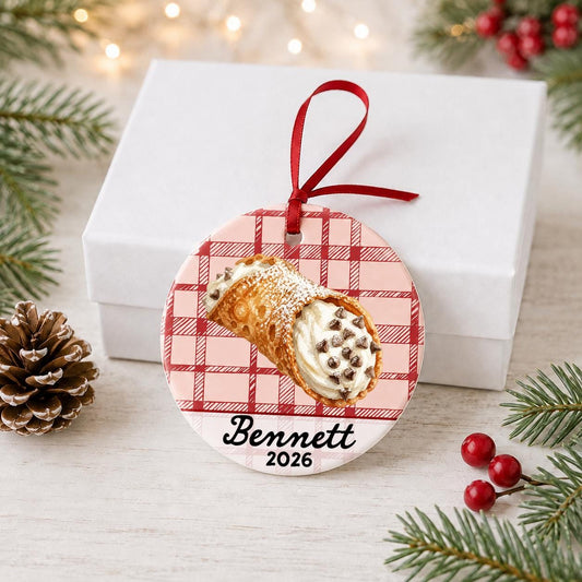 Personalized cannoli ceramic ornament with name and year on red plaid in front of a white gift box with pinecones and red berries