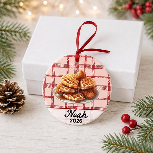 Personalized chicken and waffles ceramic ornament with name and year on red plaid in front of a white gift box with pinecones and red berries