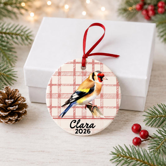 Personalized European goldfinch ceramic ornament with name and year on red plaid in front of a white gift box with pinecones and red berries