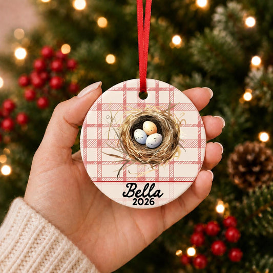 Hand holding the personalized bird nest ceramic ornament with name and year on red plaid in front of a Christmas tree