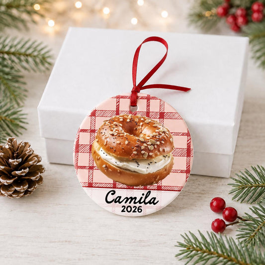 Personalized bagel ceramic ornament with name and year on red plaid in front of a white gift box with pinecones and red berries
