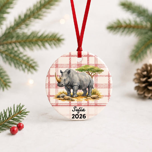 Personalized white rhinoceros ceramic ornament with name and year on red plaid with red ribbon on a white surface with pine branches and pinecones