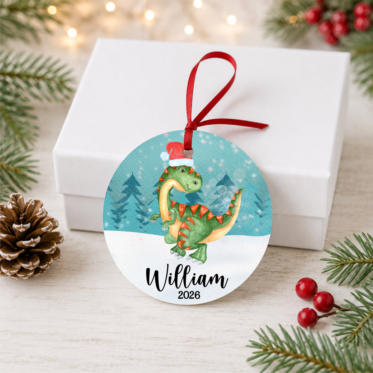 Personalized watercolor Christmas dinosaur ornament with child's name and year, displayed beside a white gift box