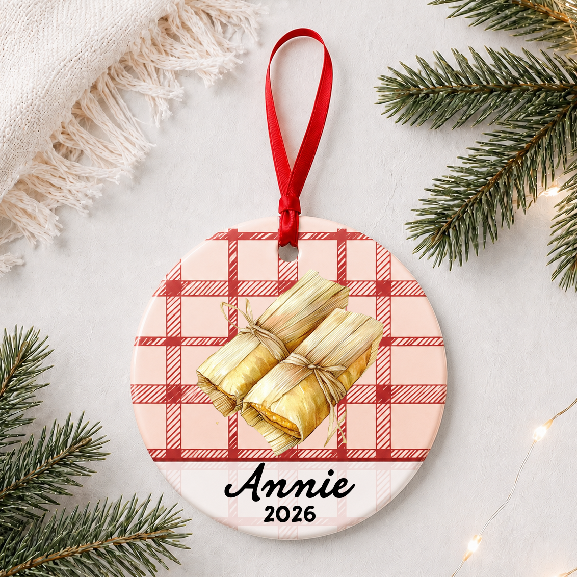 Personalized tamale ceramic ornament with name and year on red plaid styled with pine branches on a soft white background