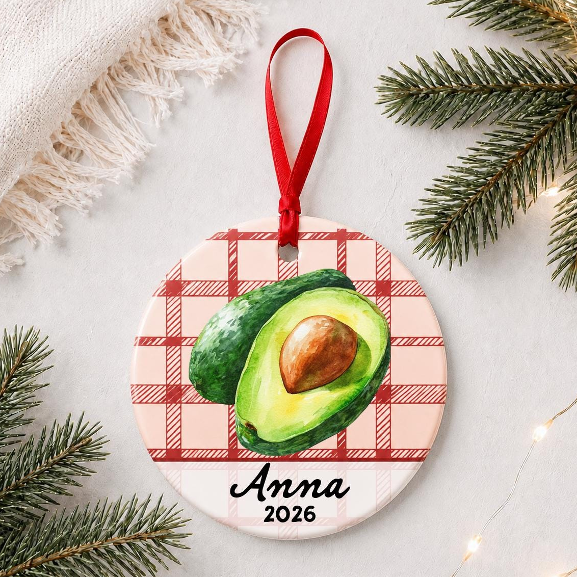 Personalized avocado ceramic ornament with name and year on red plaid styled with pine branches on a soft white background