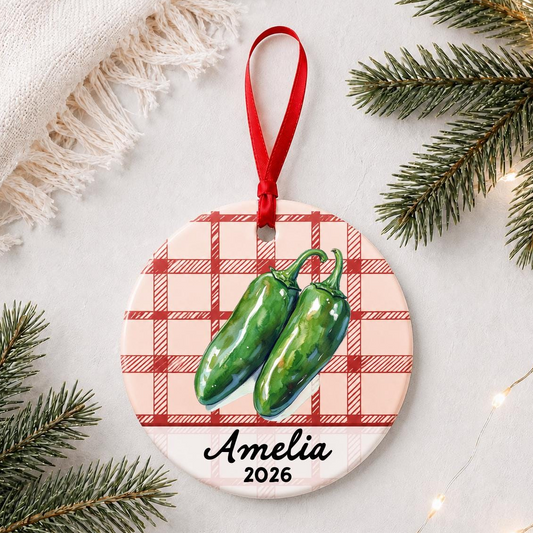 Personalized jalapeño ceramic ornament with name and year on red plaid styled with pine branches on a soft white background