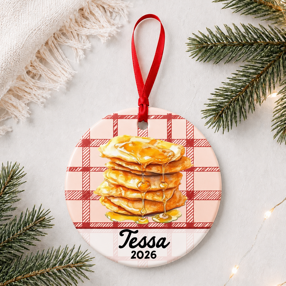 Personalized buñuelos ceramic ornament with name and year on red plaid styled with pine branches on a soft white background