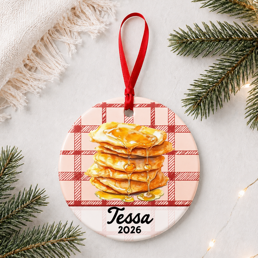 Personalized buñuelos ceramic ornament with name and year on red plaid styled with pine branches on a soft white background