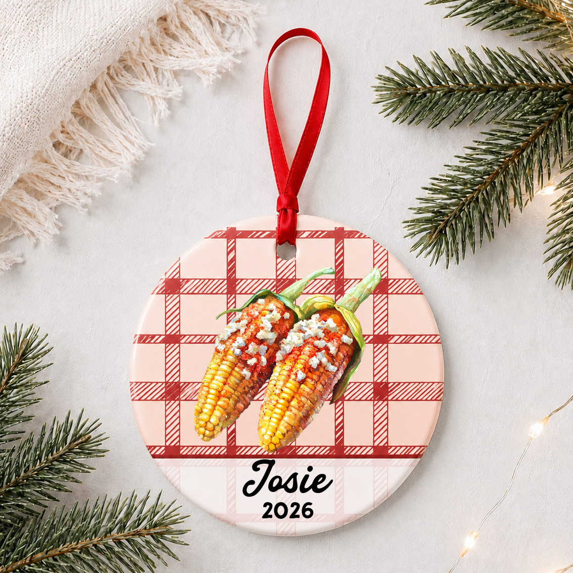 Personalized Mexican street corn ceramic ornament with name and year on red plaid styled with pine branches on a soft white background