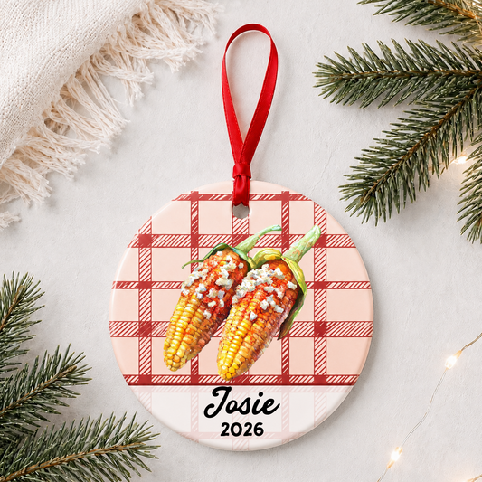 Personalized Mexican street corn ceramic ornament with name and year on red plaid styled with pine branches on a soft white background