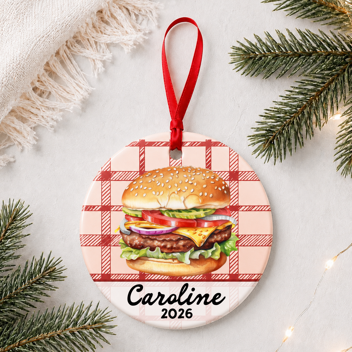 Personalized cheeseburger ceramic ornament with name and year on red plaid styled with pine branches on a soft white background
