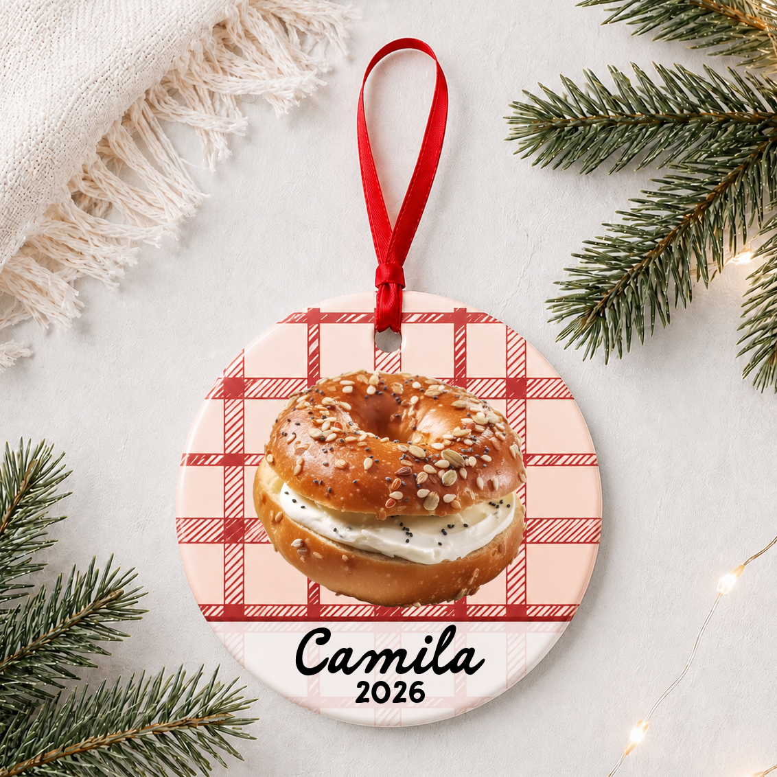 Personalized bagel ceramic ornament with name and year on red plaid styled with pine branches on a soft white background