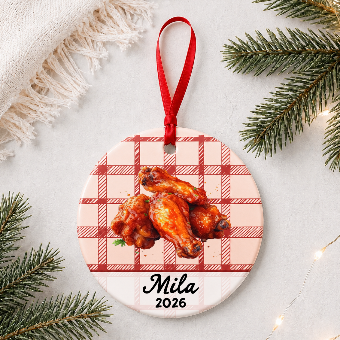 Personalized buffalo wings ceramic ornament with name and year on red plaid styled with pine branches on a soft white background