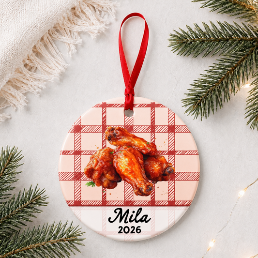Personalized buffalo wings ceramic ornament with name and year on red plaid styled with pine branches on a soft white background