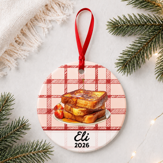 Personalized French toast ceramic ornament with name and year on red plaid styled with pine branches on a soft white background