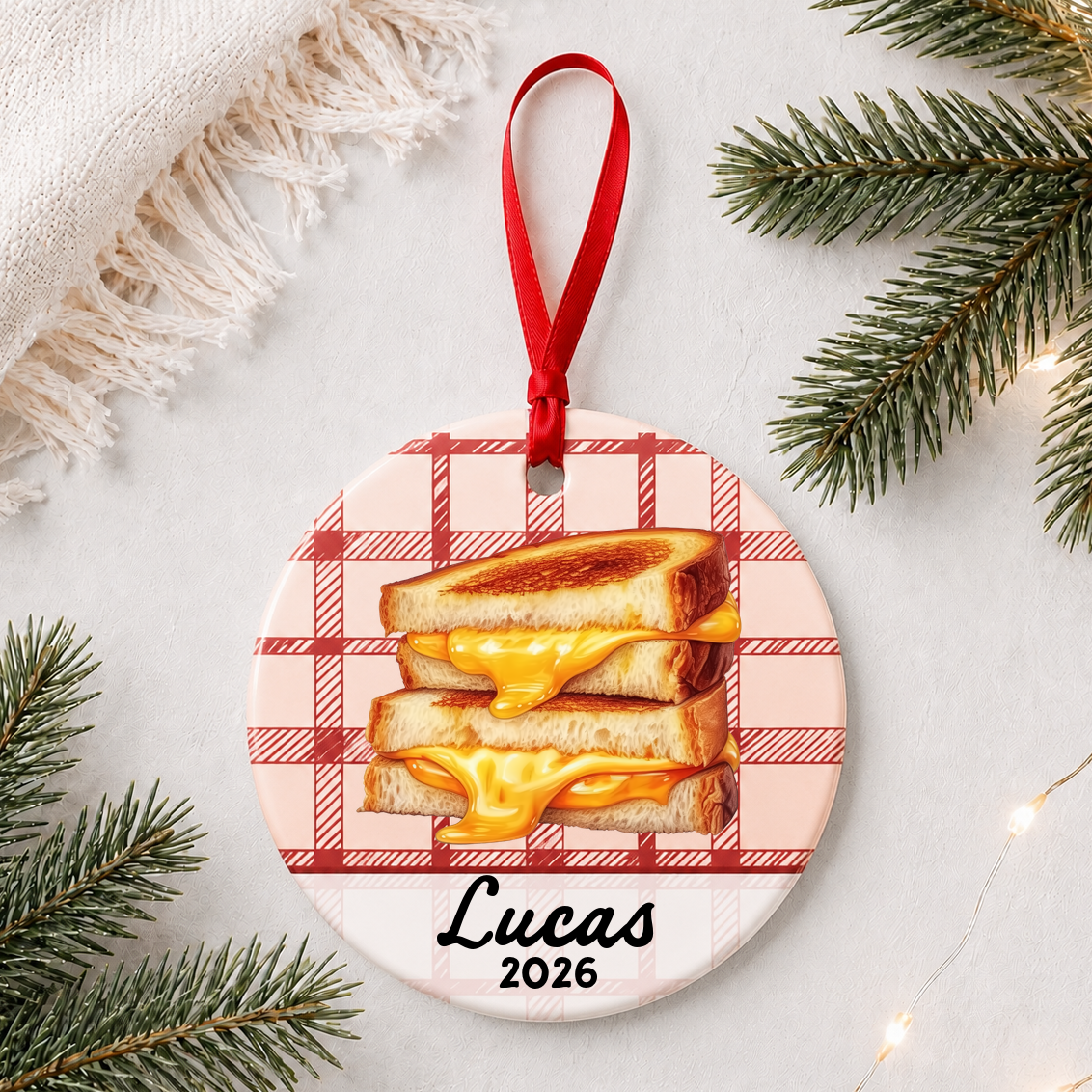 Personalized grilled cheese ceramic ornament with name and year on red plaid styled with pine branches on a soft white background
