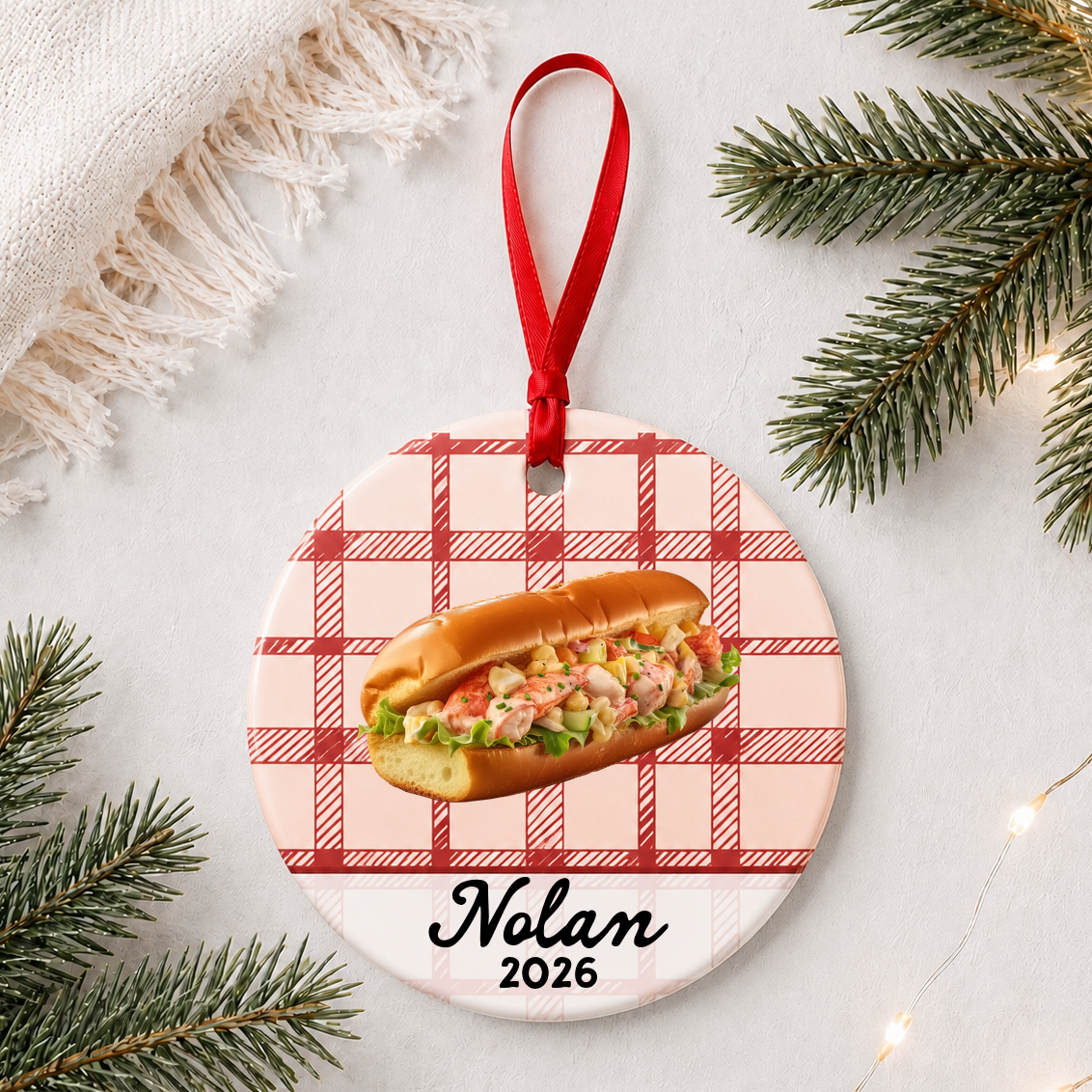 Personalized lobster roll ceramic ornament with name and year on red plaid styled with pine branches on a soft white background