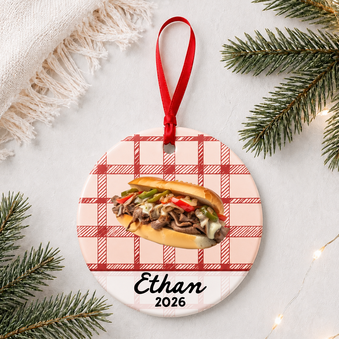 Personalized Philly cheesesteak ceramic ornament with name and year on red plaid styled with pine branches on a soft white background