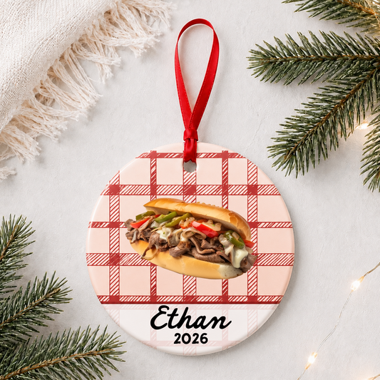 Personalized Philly cheesesteak ceramic ornament with name and year on red plaid styled with pine branches on a soft white background