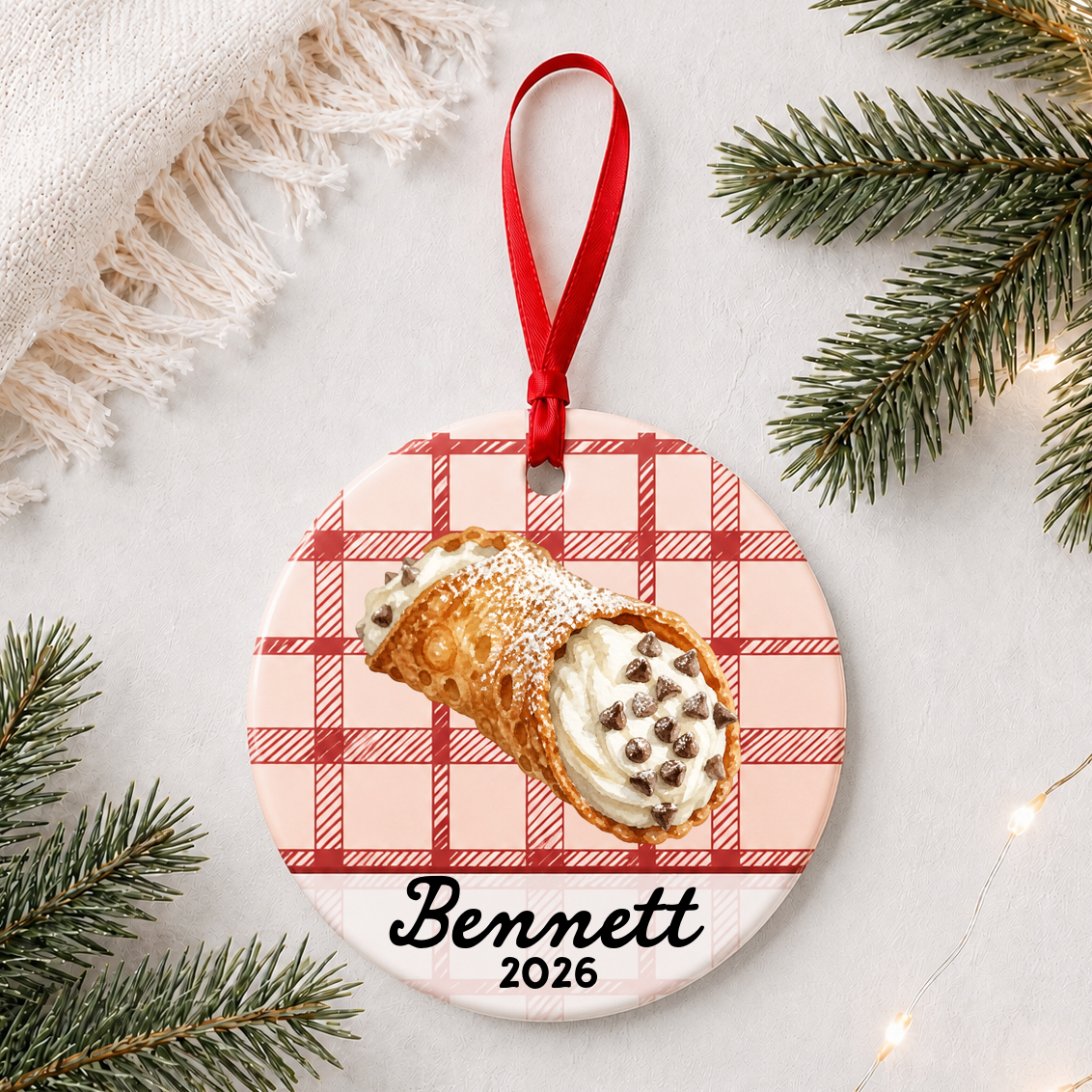 Personalized cannoli ceramic ornament with name and year on red plaid styled with pine branches on a soft white background