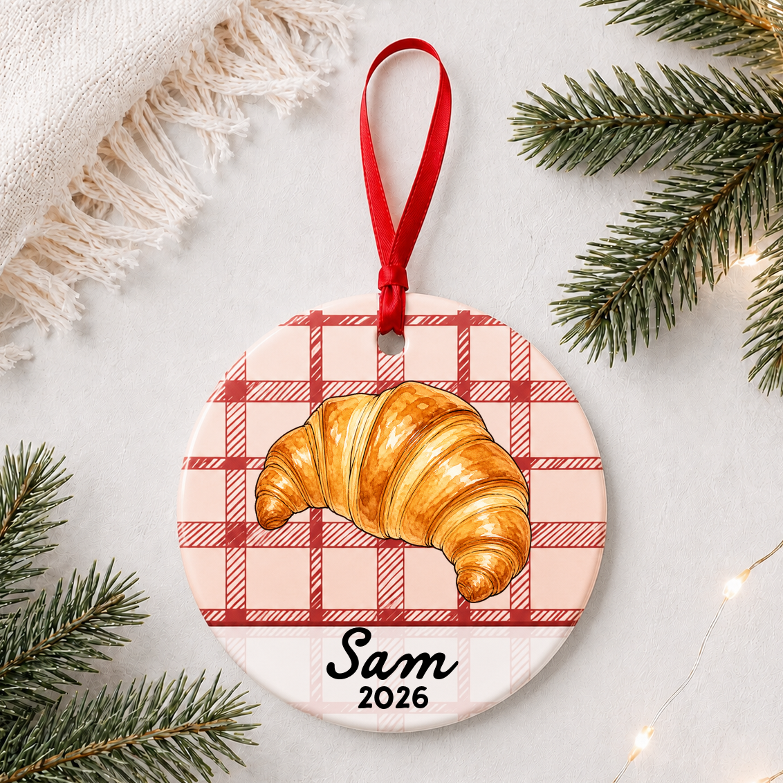 Personalized croissant ceramic ornament with name and year on red plaid styled with pine branches on a soft white background