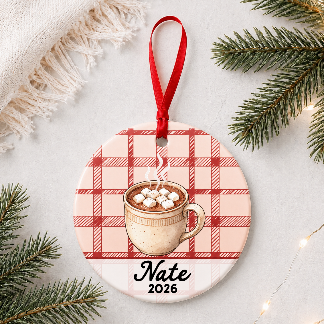 Personalized hot chocolate ceramic ornament with name and year on red plaid styled with pine branches on a soft white background