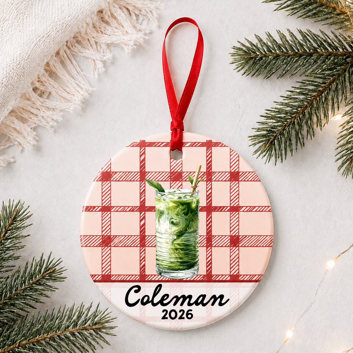 Personalized iced matcha ceramic ornament with name and year on red plaid styled with pine branches on a soft white background