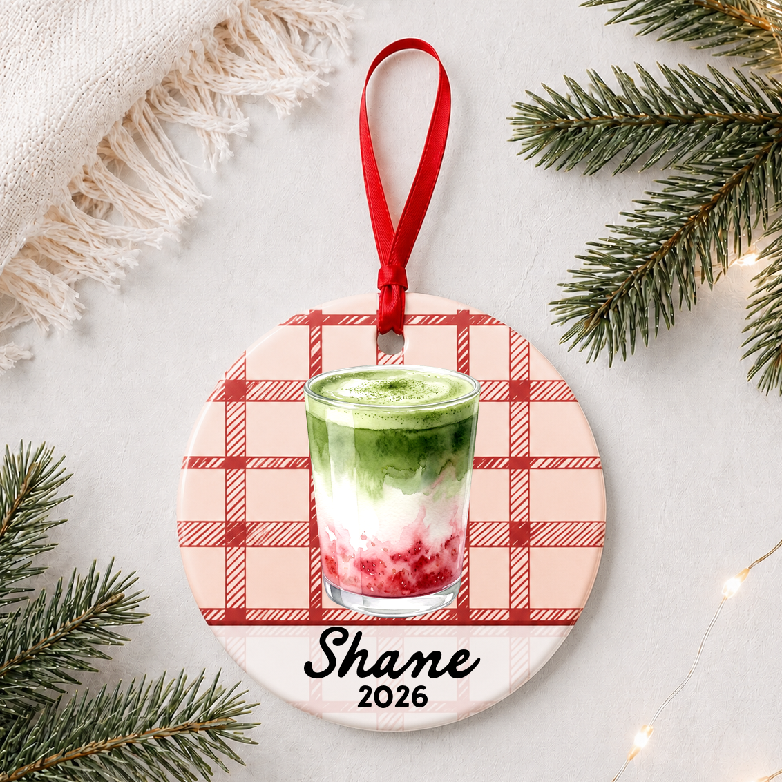 Personalized strawberry matcha latte ceramic ornament with name and year on red plaid styled with pine branches on a soft white background