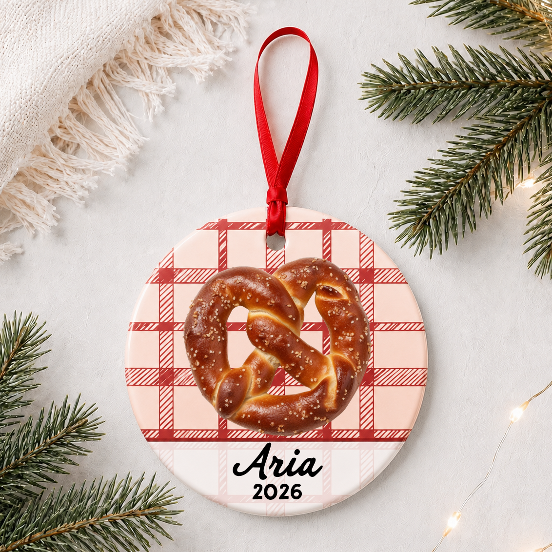 Personalized soft pretzel ceramic ornament with name and year on red plaid styled with pine branches on a soft white background