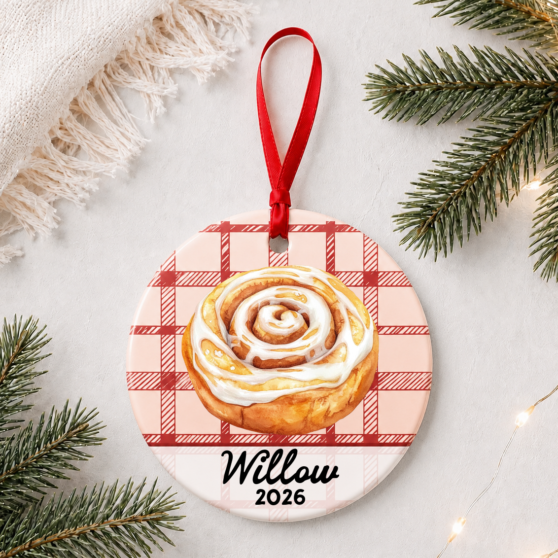 Personalized cinnamon roll ceramic ornament with name and year on red plaid styled with pine branches on a soft white background