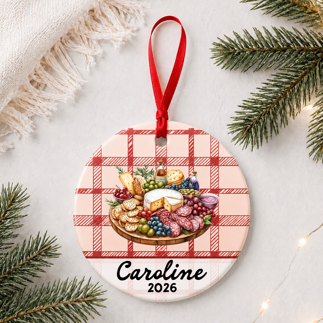 Personalized charcuterie board ceramic ornament with name and year on red plaid styled with pine branches on a soft white background