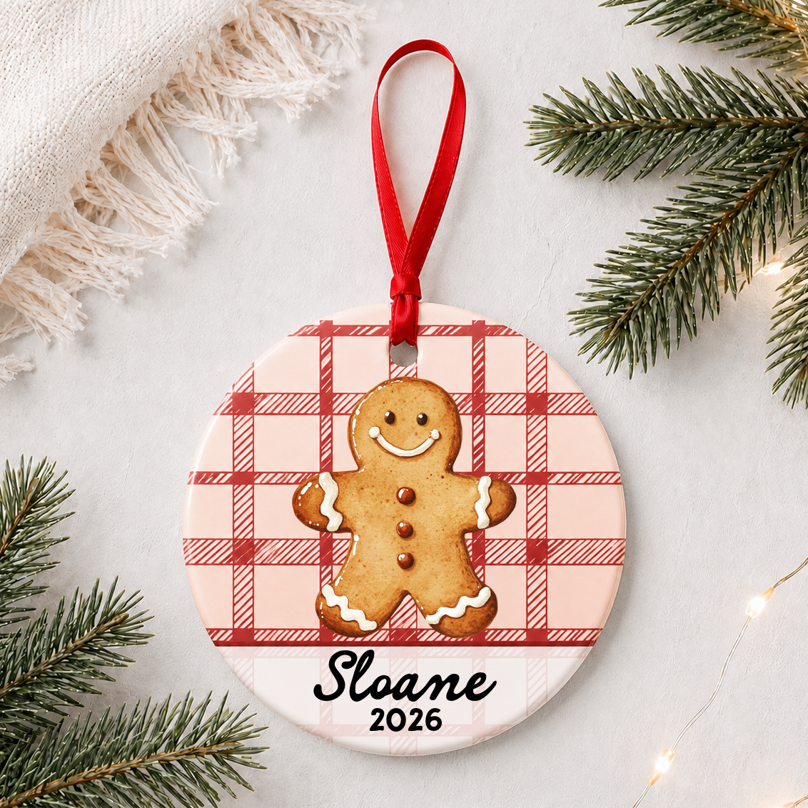 Personalized gingerbread man ceramic ornament with name and year on red plaid styled with pine branches on a soft white background