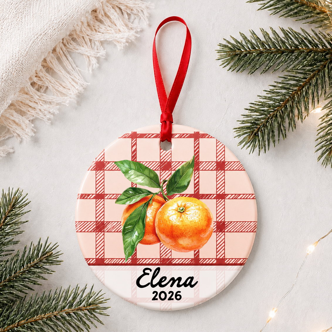 Personalized clementine ceramic ornament with name and year on red plaid styled with pine branches on a soft white background