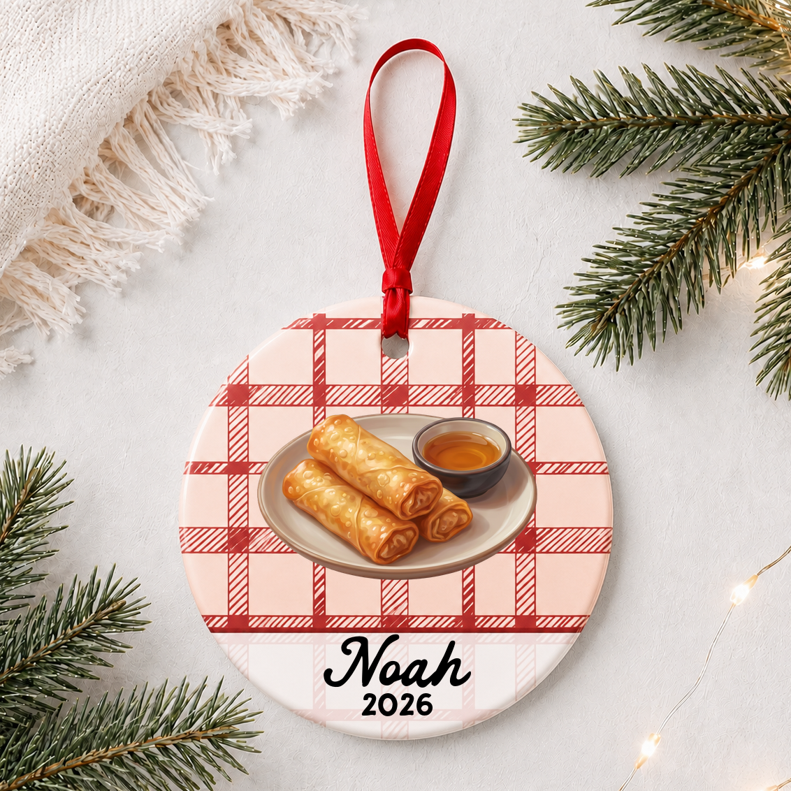 Personalized egg roll ceramic ornament with name and year on red plaid styled with pine branches on a soft white background