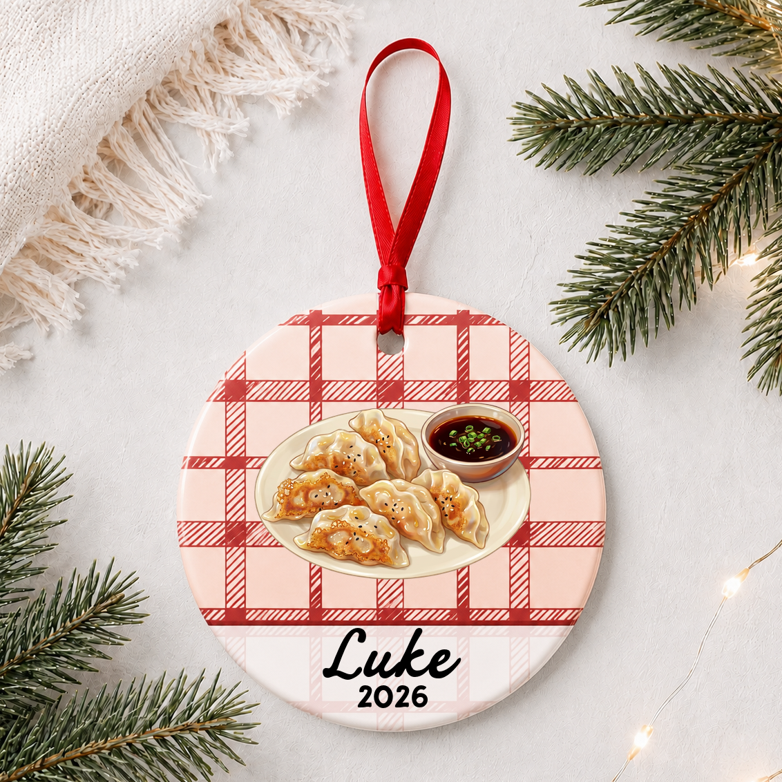 Personalized potsticker ceramic ornament with name and year on red plaid styled with pine branches on a soft white background