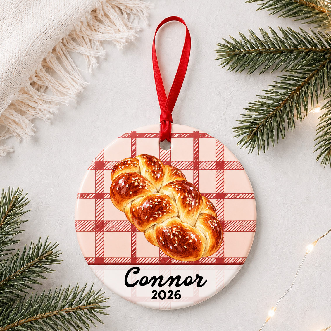 Personalized challah bread ceramic ornament with name and year on red plaid styled with pine branches on a soft white background