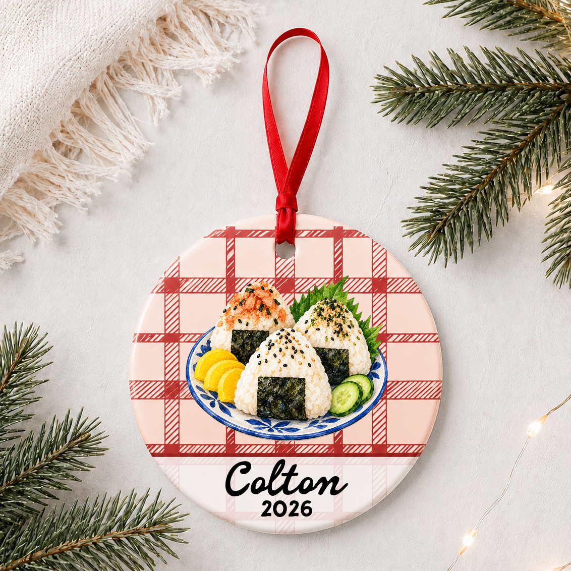 Personalized onigiri ceramic ornament with name and year on red plaid styled with pine branches on a soft white background