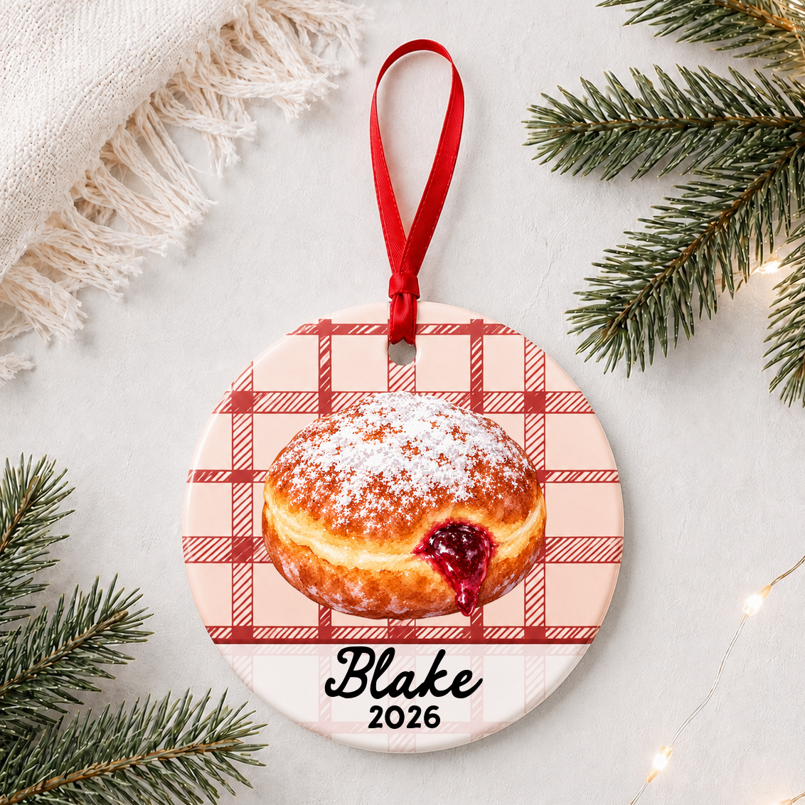Personalized paczki ceramic ornament with name and year on red plaid styled with pine branches on a soft white background