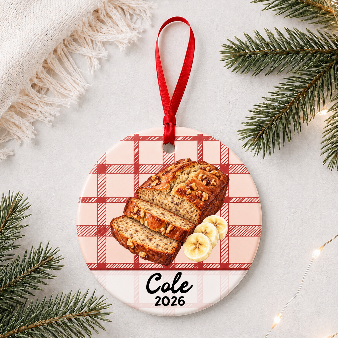 Personalized banana bread ceramic ornament with name and year on red plaid styled with pine branches on a soft white background