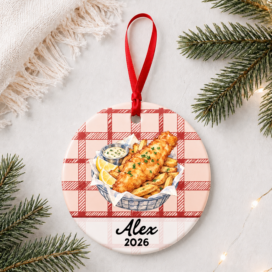 Personalized fish and chips ceramic ornament with name and year on red plaid styled with pine branches on a soft white background