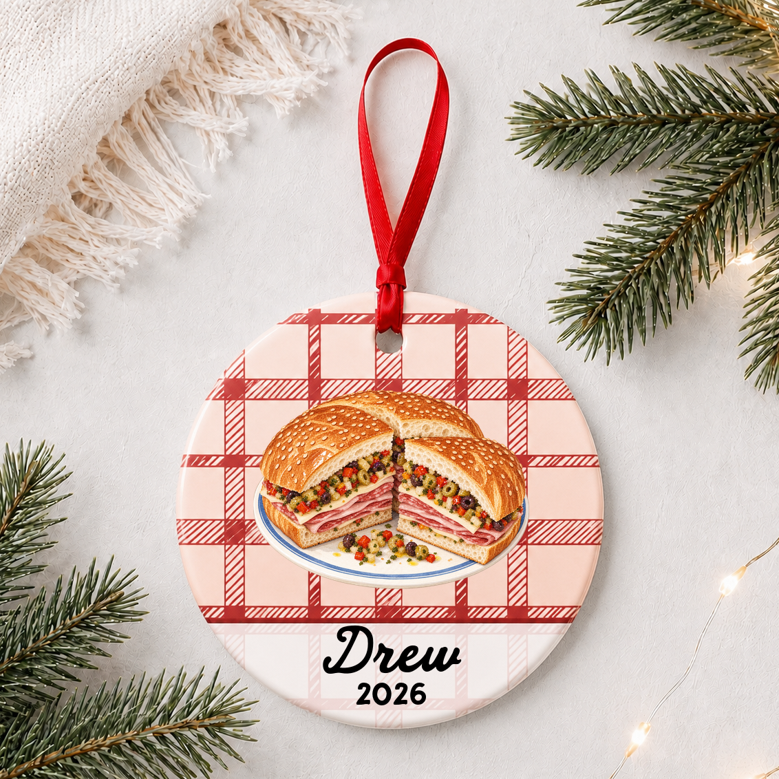 Personalized muffuletta ceramic ornament with name and year on red plaid styled with pine branches on a soft white background