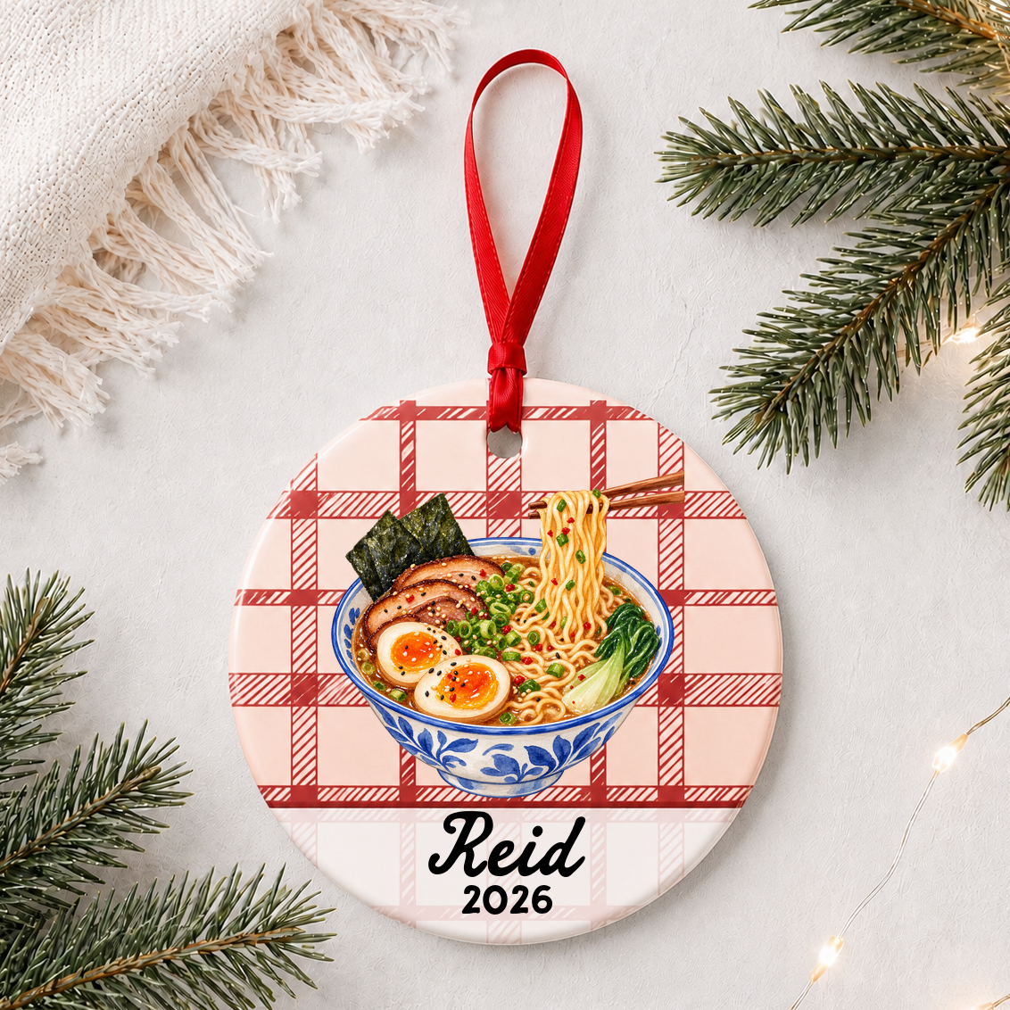 Personalized ramen ceramic ornament with name and year on red plaid styled with pine branches on a soft white background