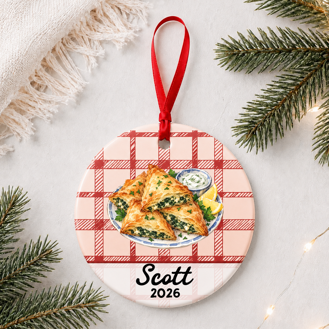 Personalized spanakopita ceramic ornament with name and year on red plaid styled with pine branches on a soft white background