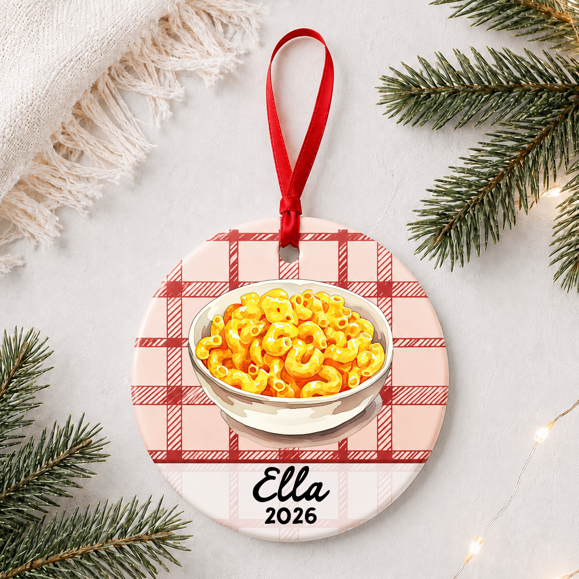 Personalized mac and cheese ceramic ornament with name and year on red plaid styled with pine branches on a soft white background
