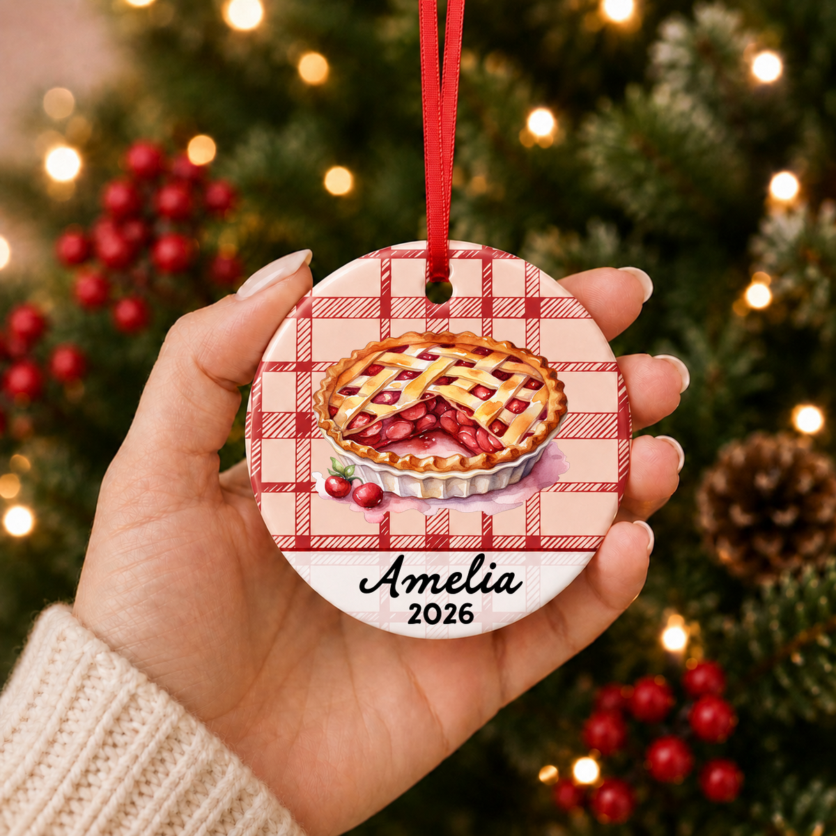 Hand holding personalized cherry pie ceramic ornament with name and year beside a Christmas tree