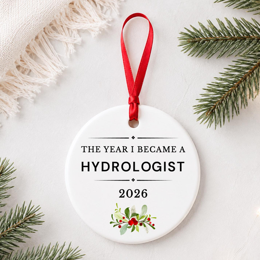 Hydrologist 2026 ceramic ornament reading The Year I Became a Hydrologist styled with pine branches on a soft white background