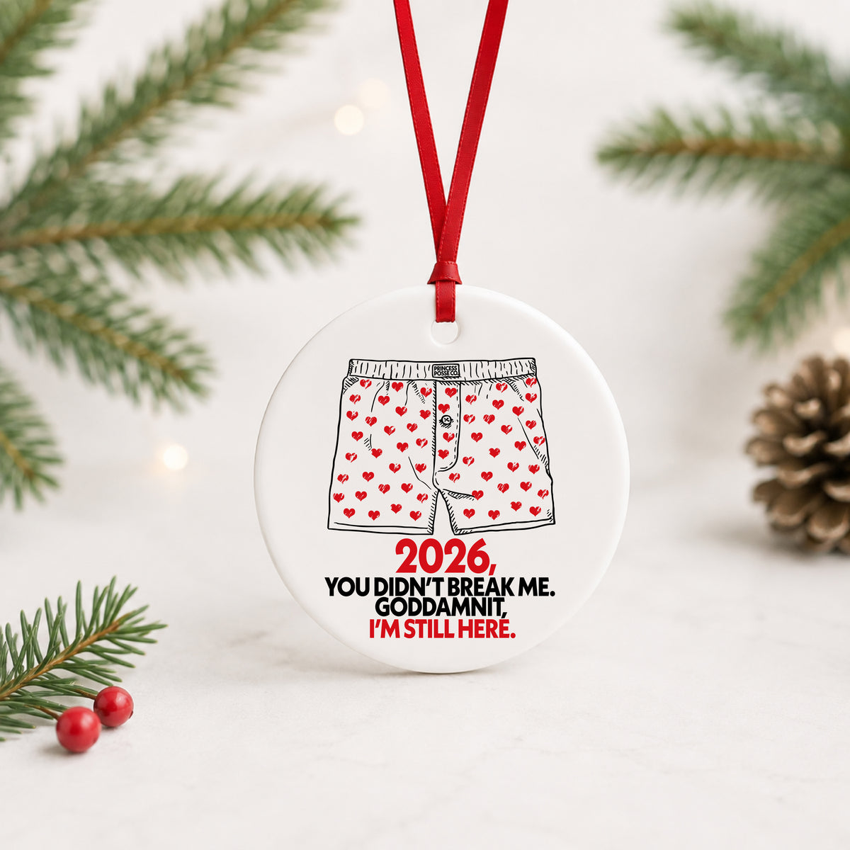 White ceramic ornament with Carl's red heart boxers and 2026 I'm still here text, red ribbon, pine branches