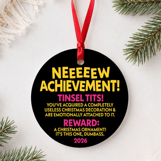 Black round Dungeon Crawler Carl New Achievement ornament with yellow and pink text and red ribbon beside pine branches