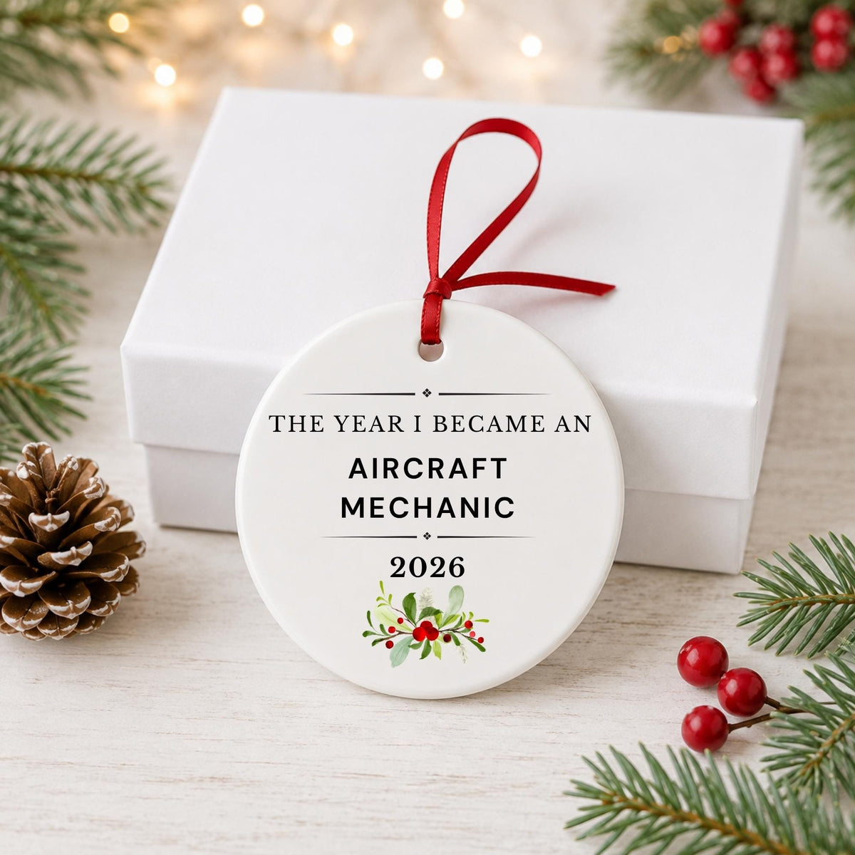 Aircraft Mechanic 2026 ceramic ornament reading The Year I Became an Aircraft Mechanic in front of a white gift box with pinecones and red berries